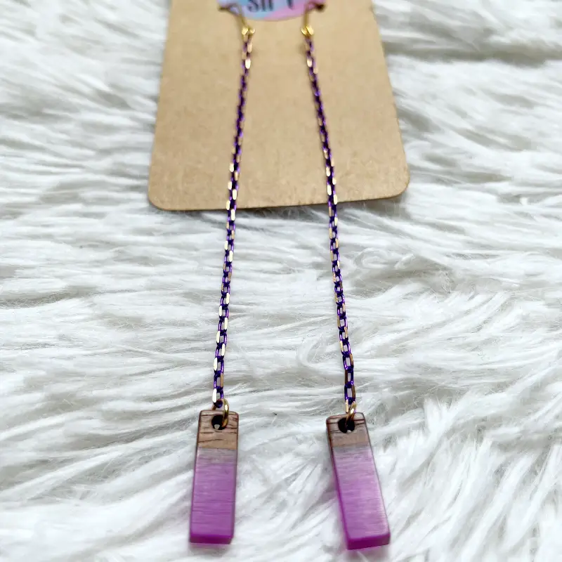 21098-violeta-4 Pendientes extra largos con cadena morada y dorada fina y una pieza rectangular pequeña de madera con resina morada.